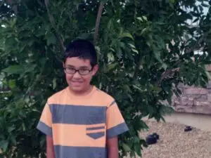 A boy in glasses and a striped t-shirt stands in front of a leafy tree and a stone wall.