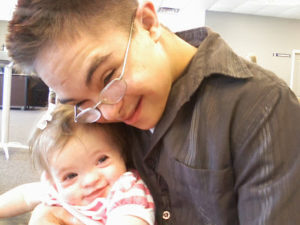 Teenage boy with Down syndrome cradling a baby girl with Down syndrome. Both with big smiles on their faces