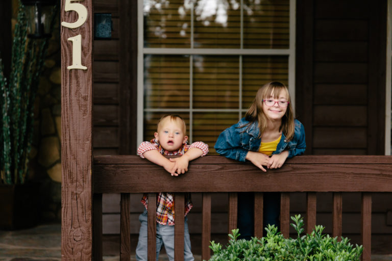 “Two children smiling while leaning over a wooden porch railing in front of a home, enjoying time together outside.”