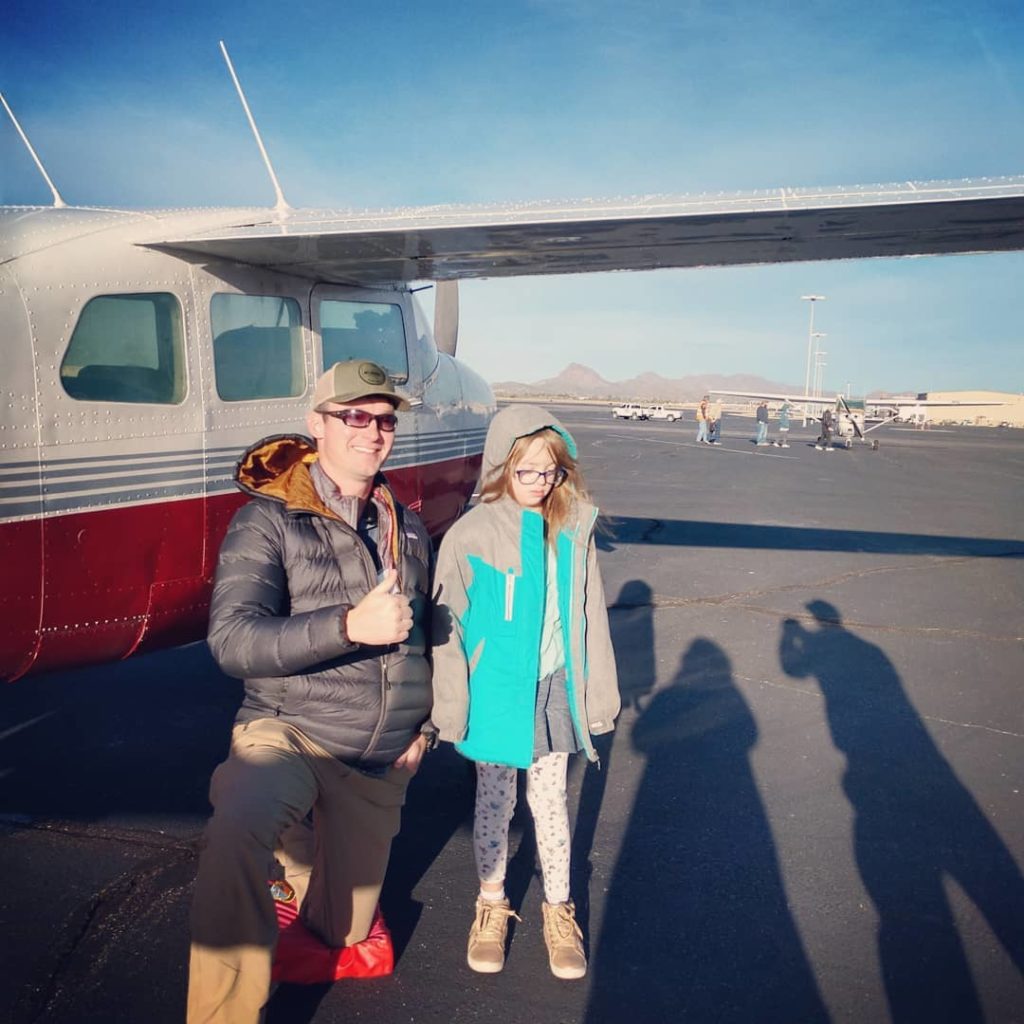 A young man kneeling on the ground next to a young girl with Down syndrome in front of an airplane.