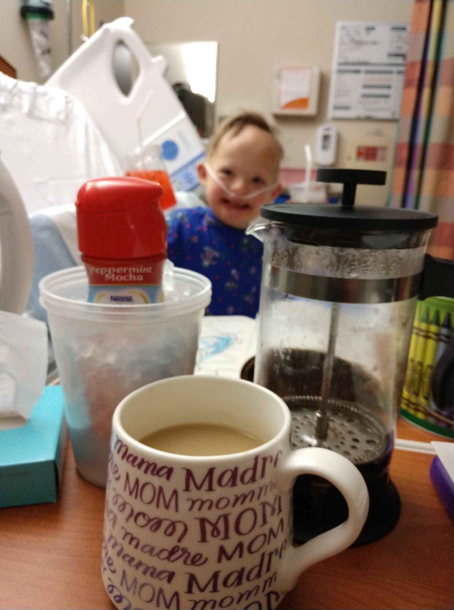 bring home to the hospital with a french press coffee maker, coffee mug, creamer sit on the table while a young boy with Down syndrome smiles in the background