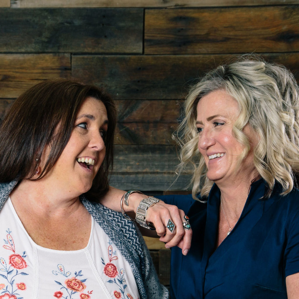 Two women laughing in front of a wooden wall, embracing the joy of life together.