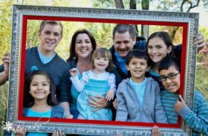 A family of eight poses outdoors, all smiling, holding a large picture frame. The children and adults are dressed in blue and teal tones.