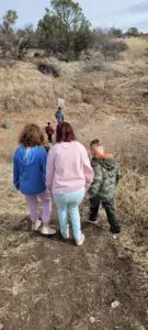 A group of people, including children and adults, walk down a dirt trail in a dry, grassy area with sparse trees.