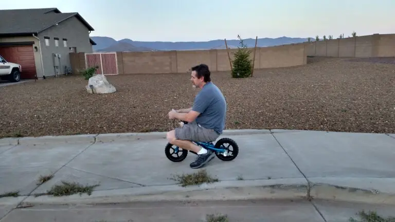 A man in a gray t-shirt and shorts rides a small child's bicycle on a sidewalk in a residential area with a house, a rock, and a distant mountain range in the background.