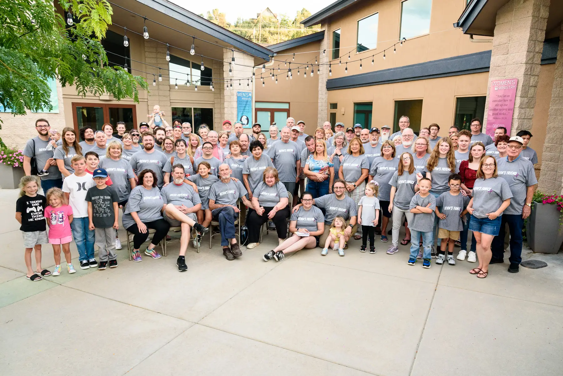 A large group of people, wearing matching gray T-shirts, gather for a group photo in an outdoor courtyard with string lights overhead and buildings in the background. Some are sitting, others are standing.