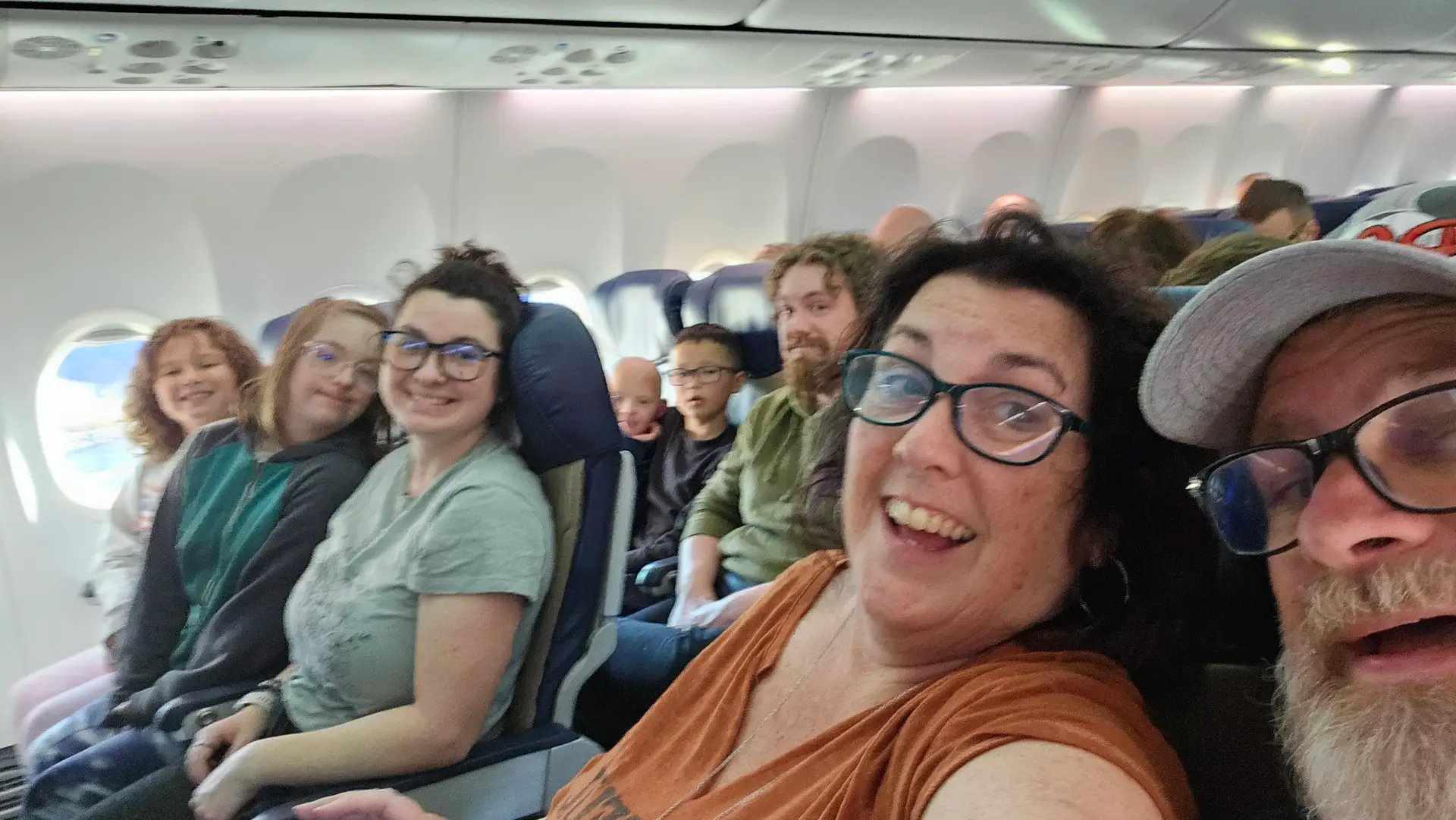 A group of smiling people, including children and adults, are seated side by side inside an airplane.