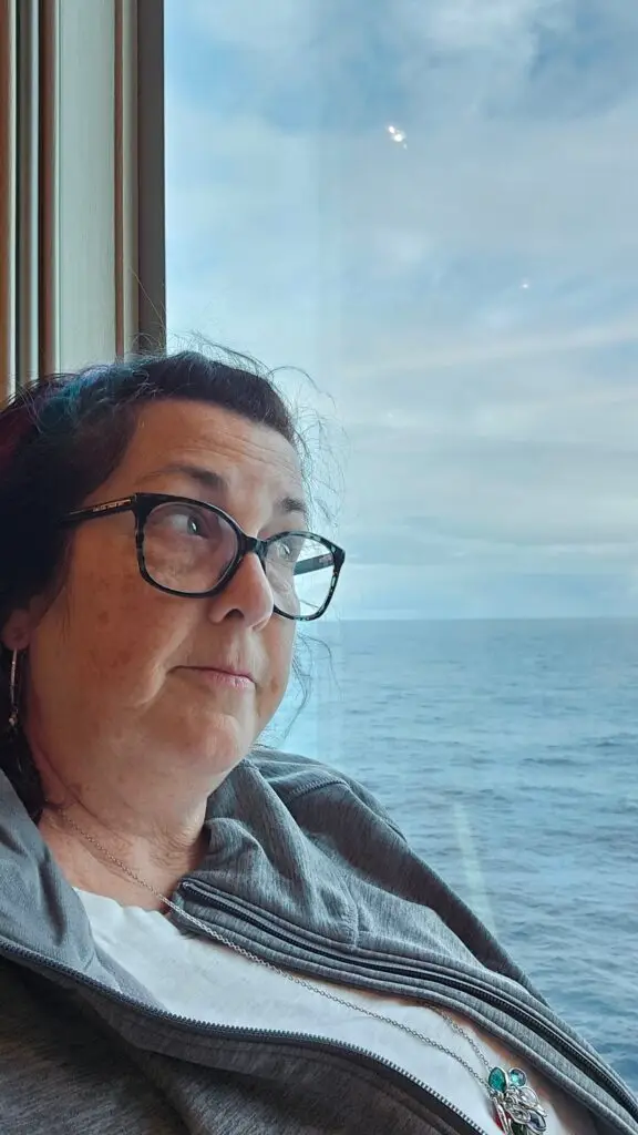 A woman wearing glasses wisitting by a cruise ship window gazing out to the ocean