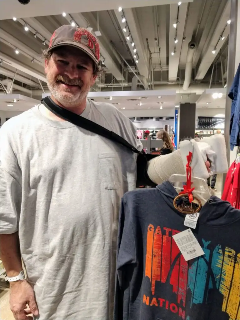 Person smiling and standing next to a clothing display in a store, wearing a grey shirt and baseball cap. The clothing display features a hoodie with the text "Nation." on a hanger hanging from his cast