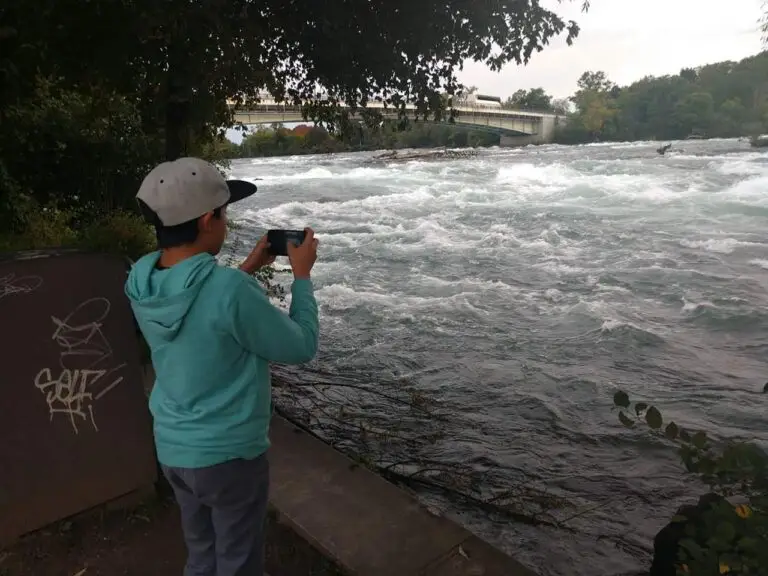 A person wearing a hoodie and cap is taking a photo with a smartphone near a rapidly flowing river, with a bridge visible in the background stretching over the Niagara Falls