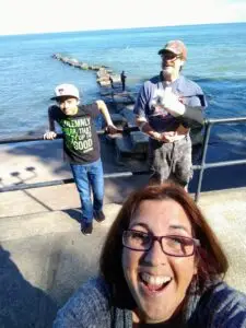 A woman takes a selfie while smiling. In the background, a man with his arm in a sling and a young boy wearing a cap stand near a railing by a body of water with a rocky jetty.