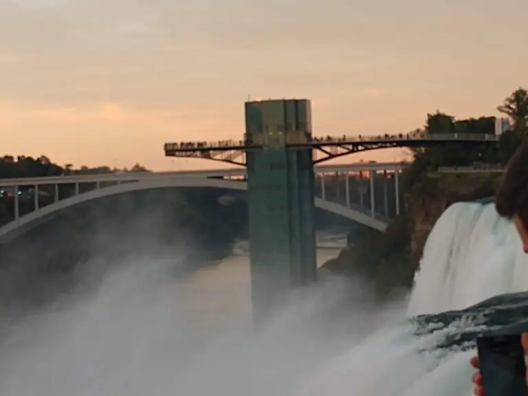 Observation deck and bridge at Niagara Falls during sunset, viewed from the edge of the waterfall with mist rising from the falls. People are visible on the observation platform and taking photos.