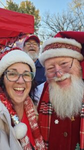 A woman and two men wearing festive Santa hats smile outdoors on a sunny day, with a red canopy and trees in the background.