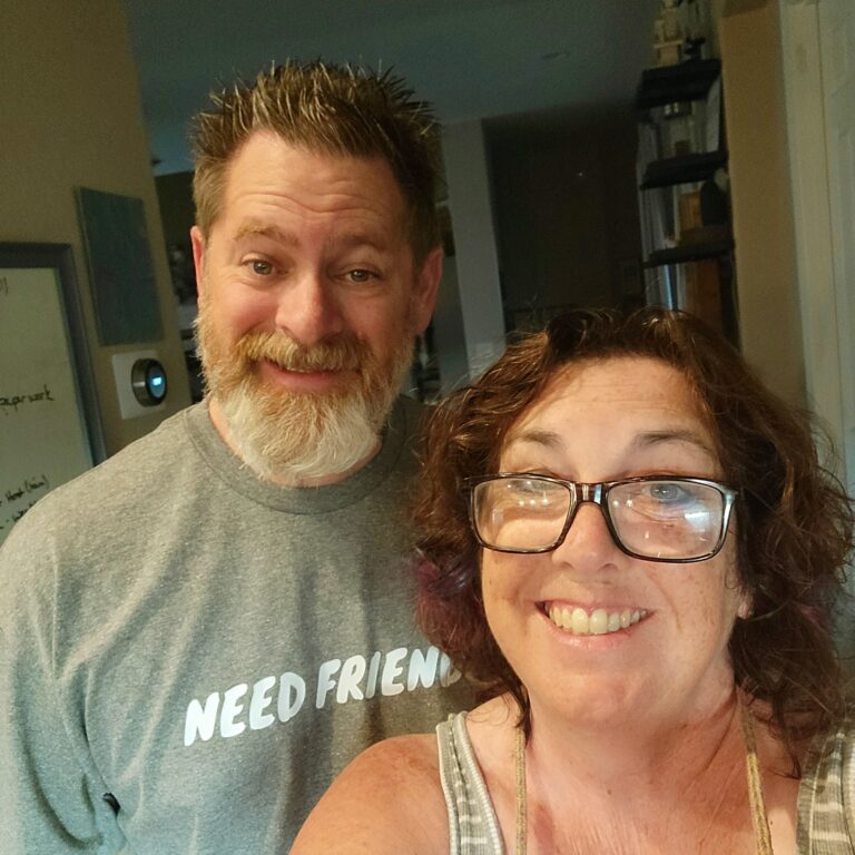 Kevin sits at the kitchen counter wearing a “Need Friends” shirt, smiling softly as sunlight filters through the window. The shirt reflects his family’s hope that gentle social engagement will help slow the progression of his Alzheimer’s.