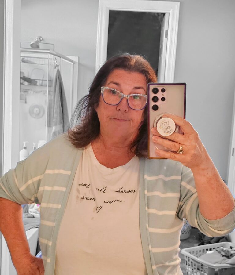 Woman in glasses and a “Not all heroes wear capes” shirt taking a mirror selfie, symbolizing everyday faith and purpose.