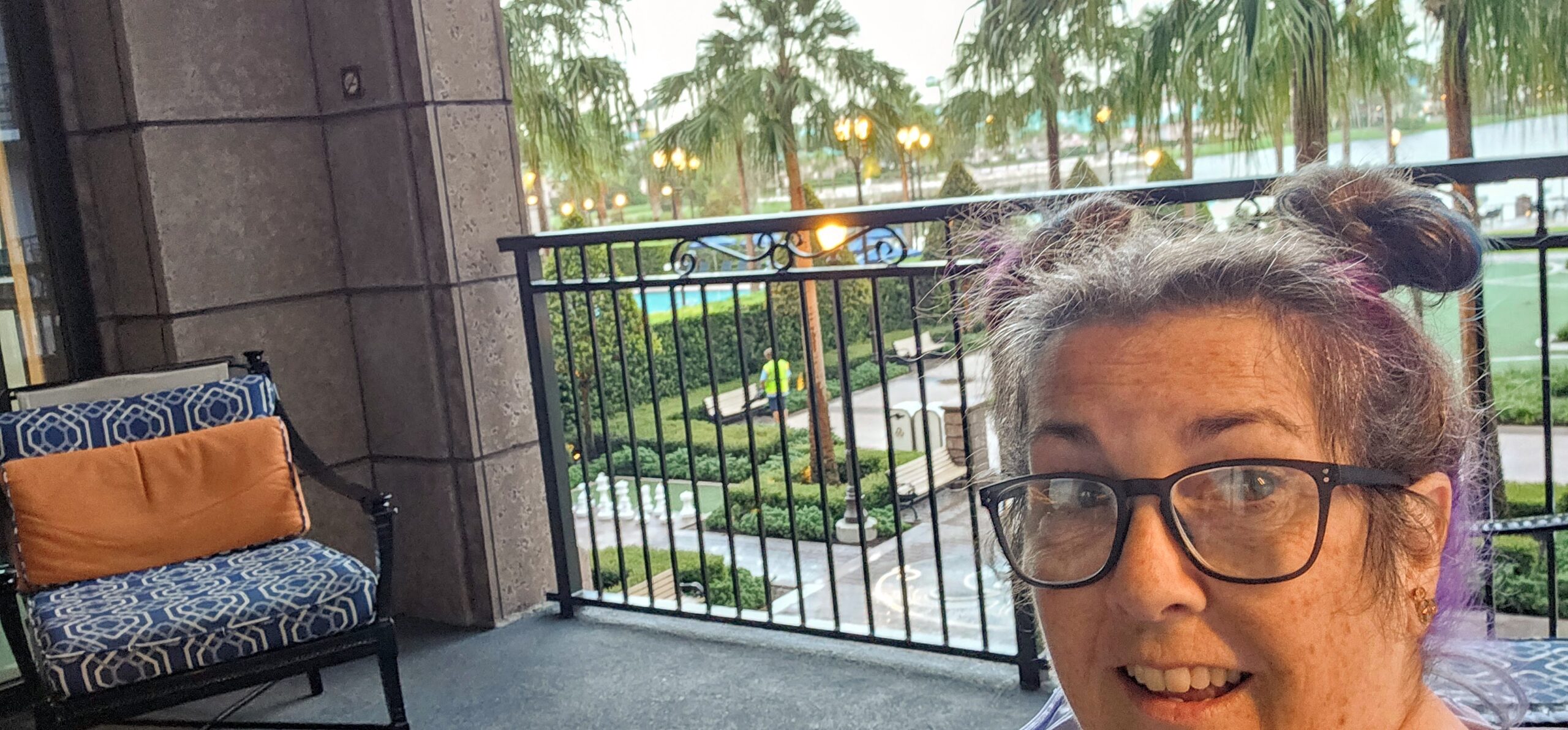 A woman with glasses and a playful double bun hairstyle smiles while enjoying a quiet Disney morning on a balcony overlooking palm trees and gardens.