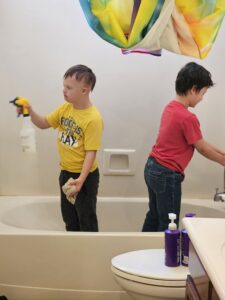 “Two children standing in a bathtub while cleaning; one sprays cleaner and the other wipes the faucet as part of family bathroom chores.”