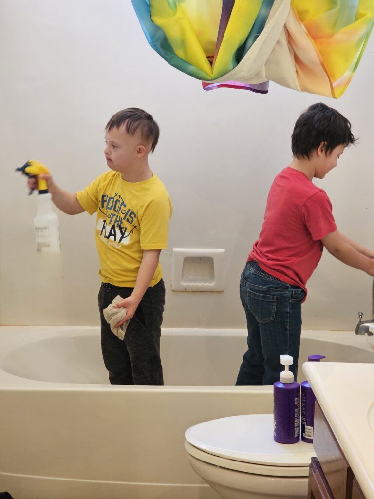 “Two children standing in a bathtub while cleaning; one sprays cleaner and the other wipes the faucet as part of family bathroom chores.”