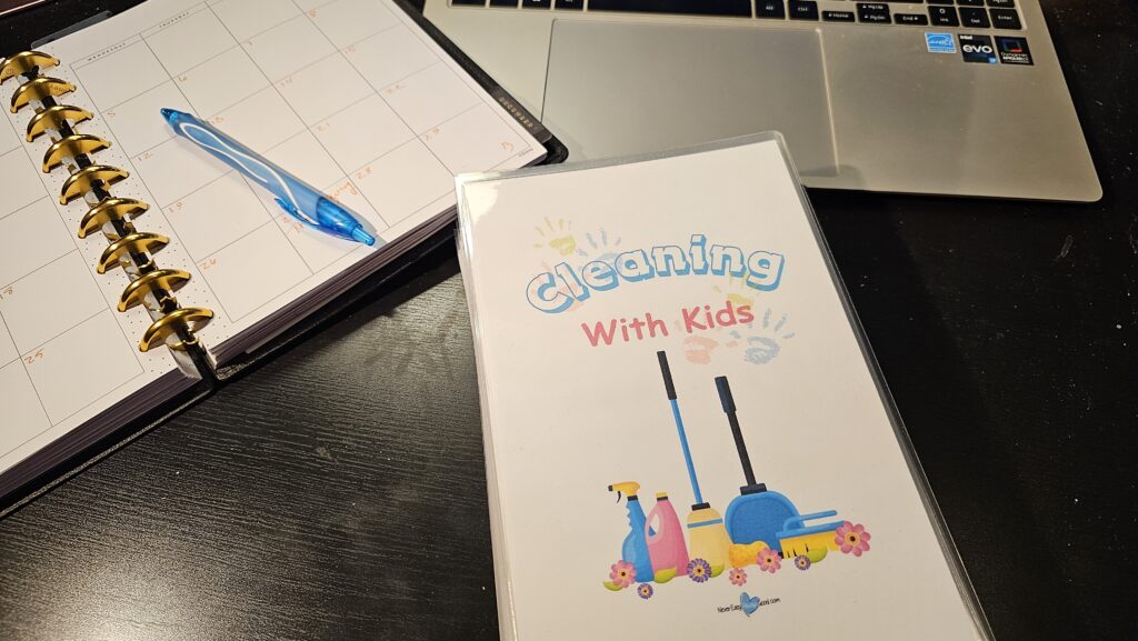 “Cleaning With Kids booklet on a desk next to an open calendar planner, blue pen, and laptop.”