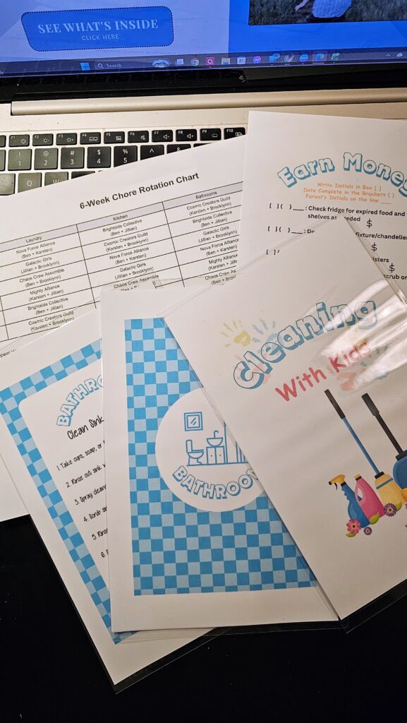 Printed Cleaning With Kids chore cards, bathroom task sheets, and a 6-week chore rotation chart laid out on a laptop, showing tools used to organize family responsibilities.