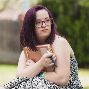 A teenage girl with Down syndrome sits outside holding a spiral notebook and pen, her purple hair shining in the sunlight. She looks thoughtful and creative, wearing red glasses and colorful wristbands.