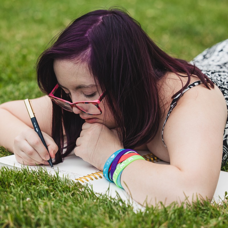 A teenage girl with Down syndrome lies on the grass writing in a notebook. She has dark purple hair, pink glasses, and colorful wristbands, focused thoughtfully on her writing.