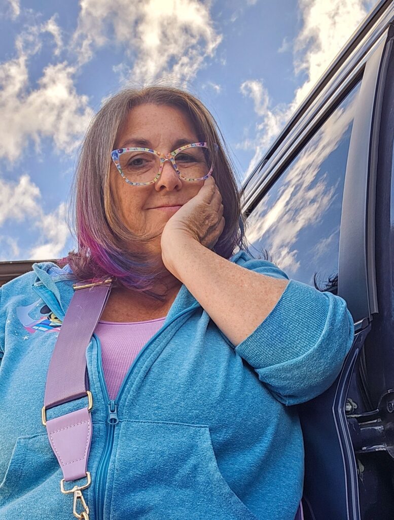 Woman with colorful glasses and purple-streaked hair wearing a blue hoodie and lavender strap bag, smiling softly beside a car under a bright blue sky with clouds.