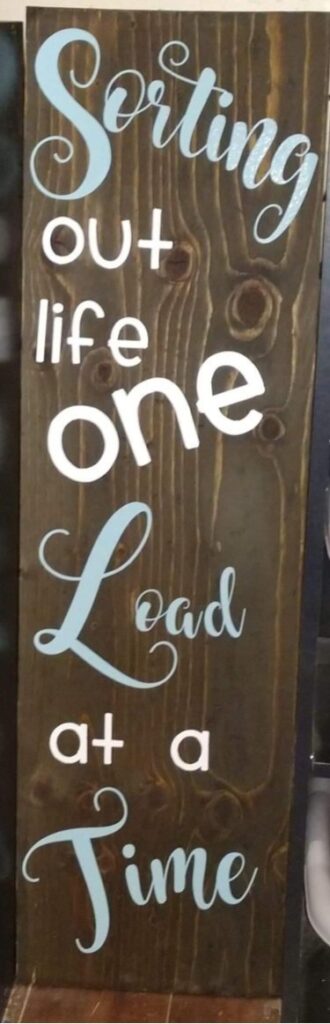 Wooden laundry room sign with the words “Sorting out life one load at a time” painted in blue and white script.