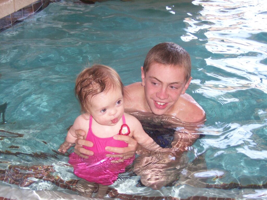 An older sibling holding a baby in a swimming pool, showing care, trust, and family support during a season of growth and transition.